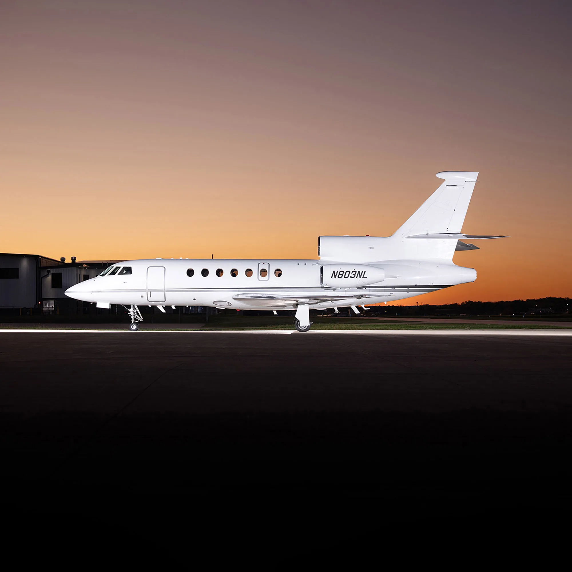 A white Dassault Falcon 900EX business jet (N803NL) parked on an airfield at sunset.