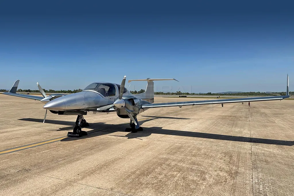 A silver and dark grey Diamond DA62 aircraft parked on an asphalt runway under a clear blue sky.