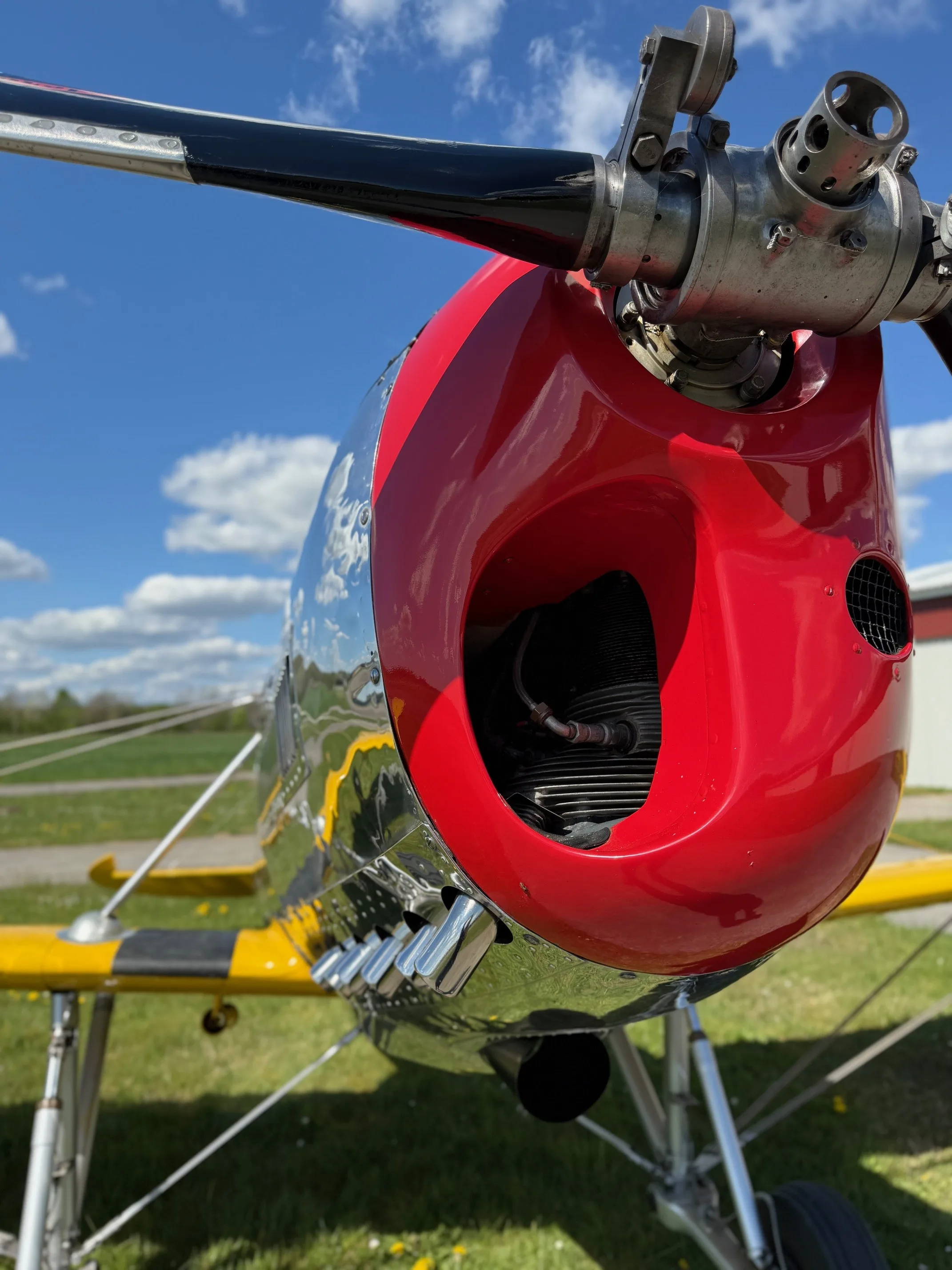 A close-up view of the red and chrome engine cowling and propeller of a vintage aircraft, with a blue sky and clouds in the background.