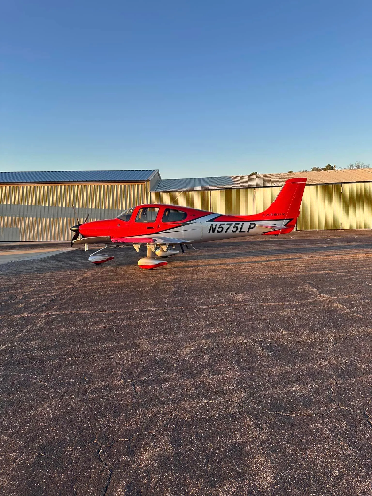 A red and silver Cirrus SR22 aircraft (N575LP) parked on an asphalt ramp with hangars in the background during golden hour.
