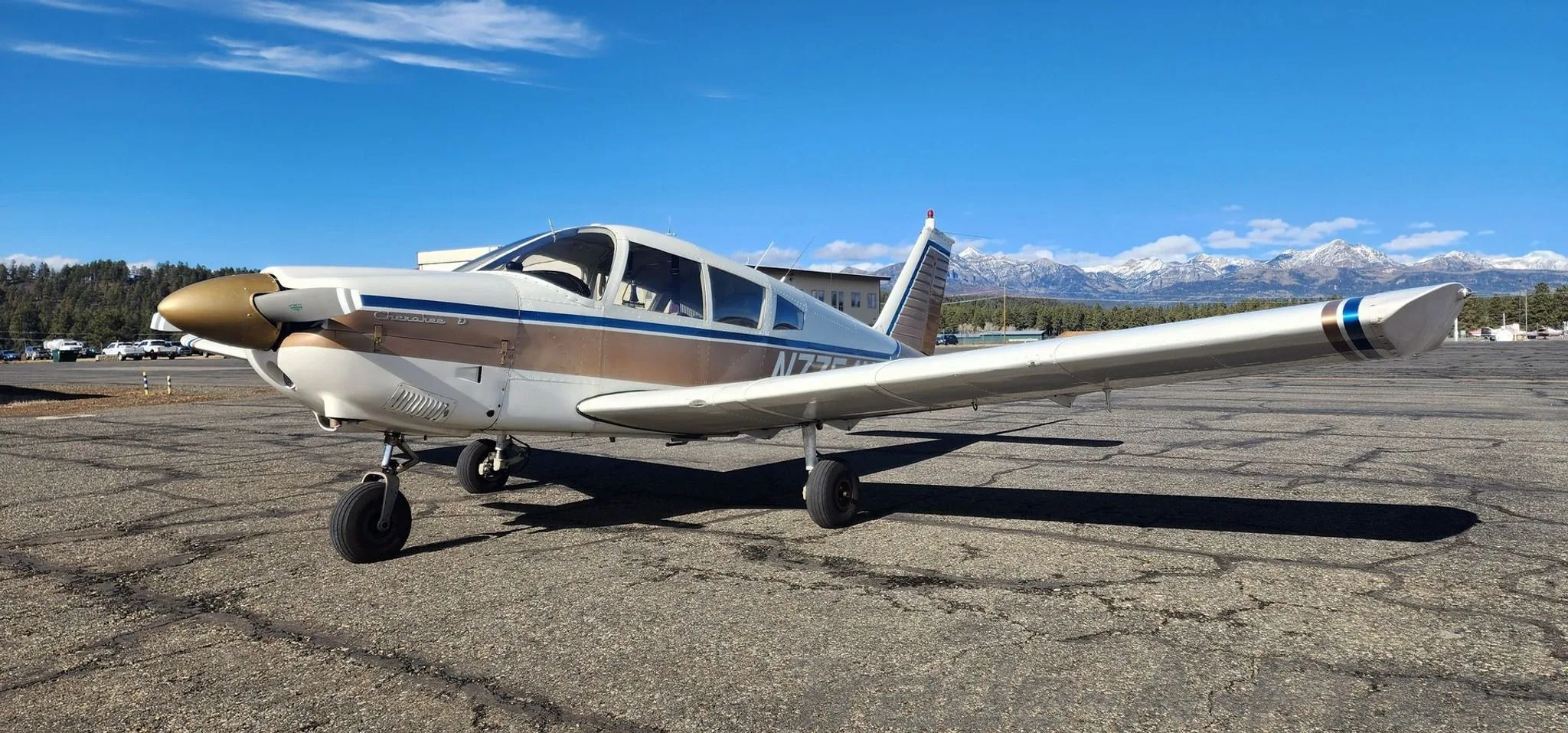 A front-left view of a white and gold Piper Cherokee 140 (N7754N) on an airfield with mountains in the distance.