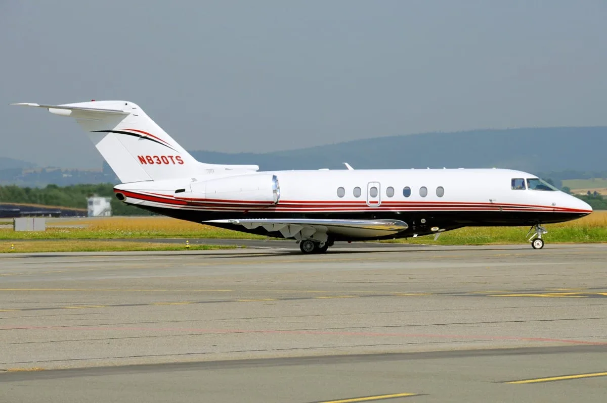 A white business jet with red and black stripes (N830TS) is parked on an airport tarmac.