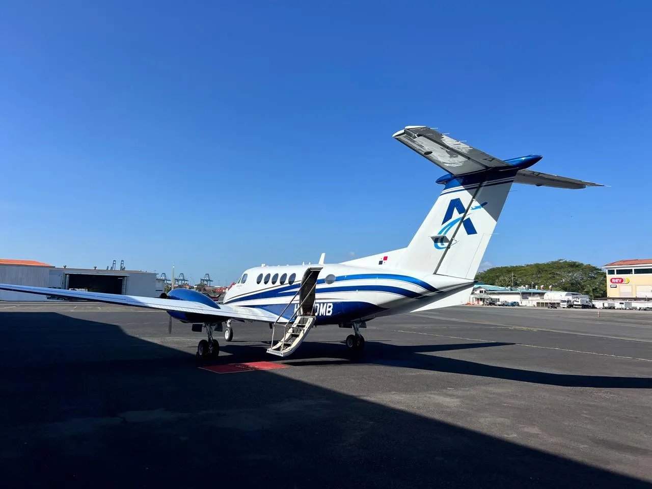 A white and blue turboprop aircraft (King Air) with registration HP-5DMB and its airstair door open, parked on an airport tarmac under a clear blue sky.