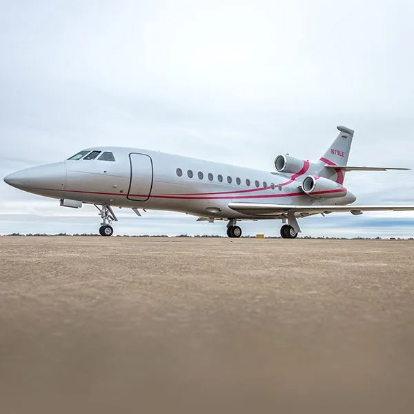 A silver Dassault Falcon 900EX private jet with pink stripes on a tarmac under a cloudy sky.