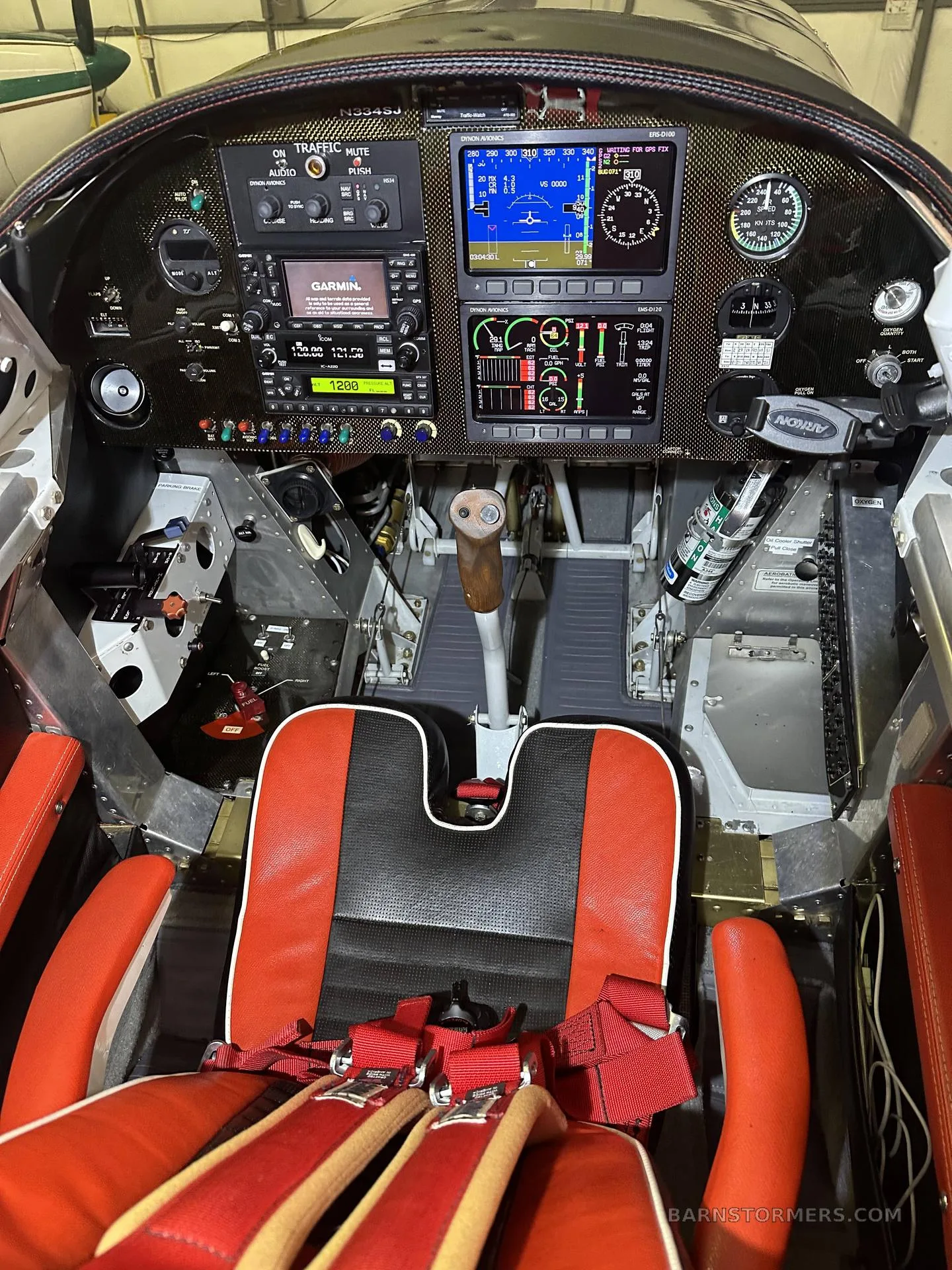 The cockpit of an aircraft with a modern instrument panel, red and black seats, and a central control stick.