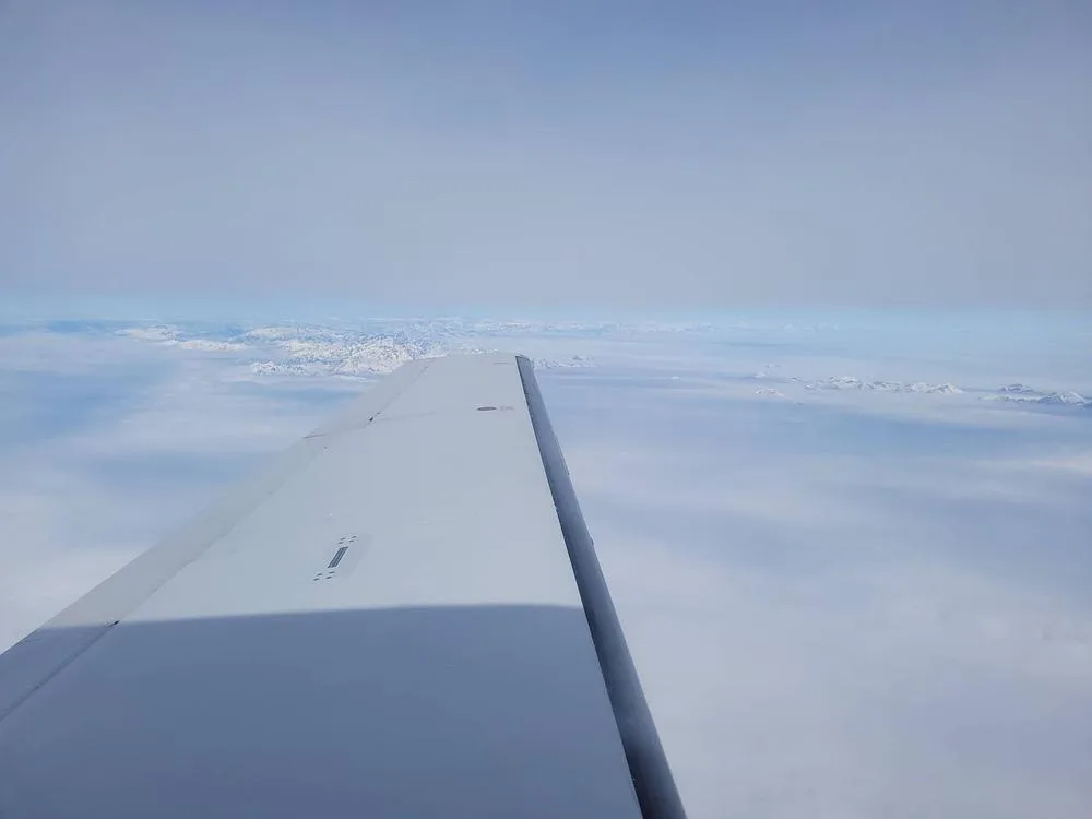 The white wing of a Piper PA-46-350P Malibu Mirage aircraft flying above clouds with distant mountains visible on the horizon.