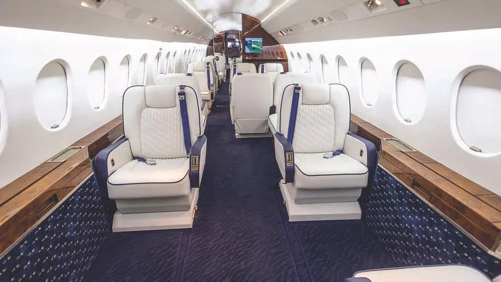 The aircraft cabin interior features white quilted seats with blue accents and a dark blue patterned carpet.