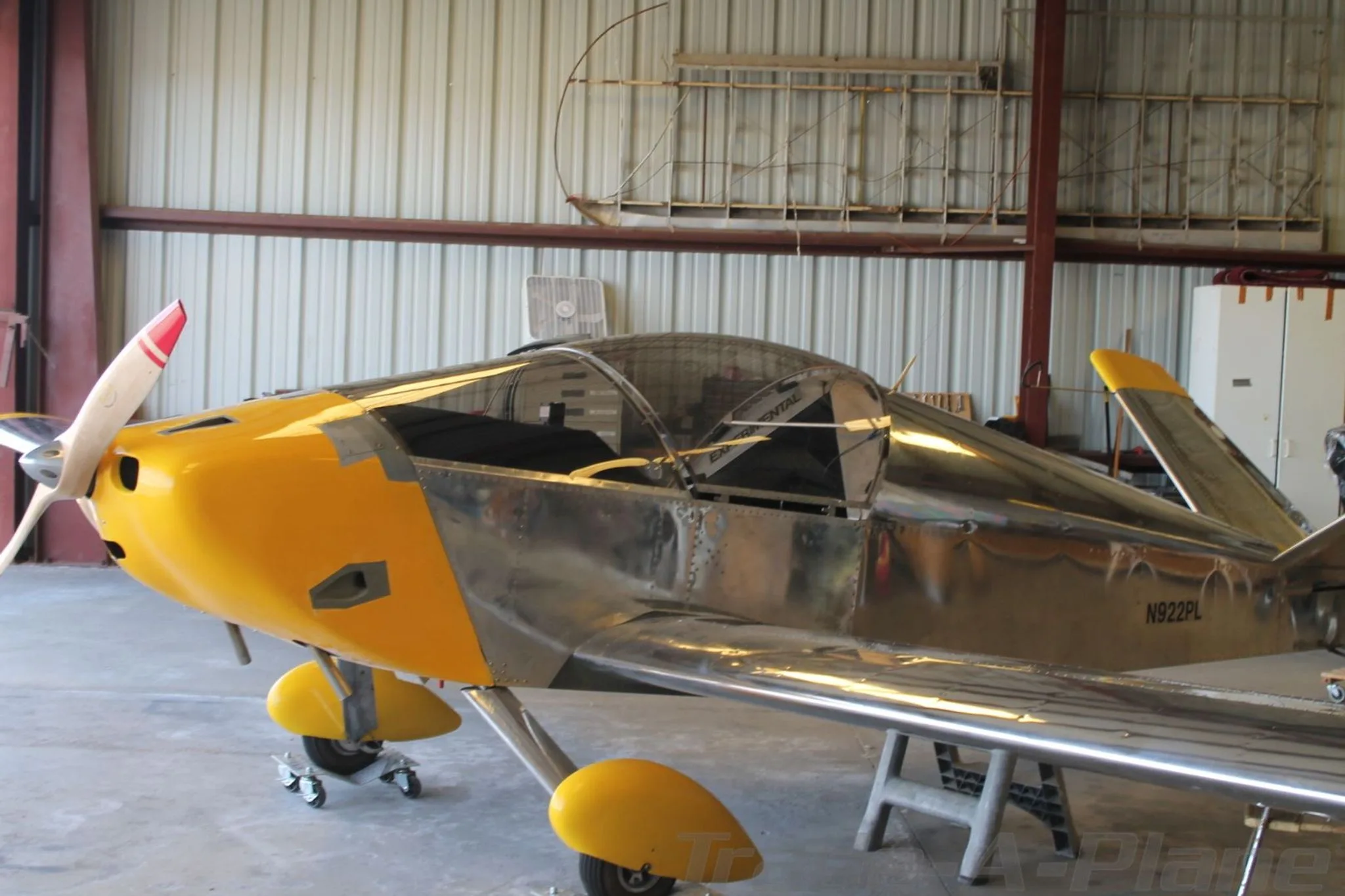 A partially assembled, unpainted metal aircraft with a yellow nose and tail number N922PL, viewed from the front-left in a hangar.