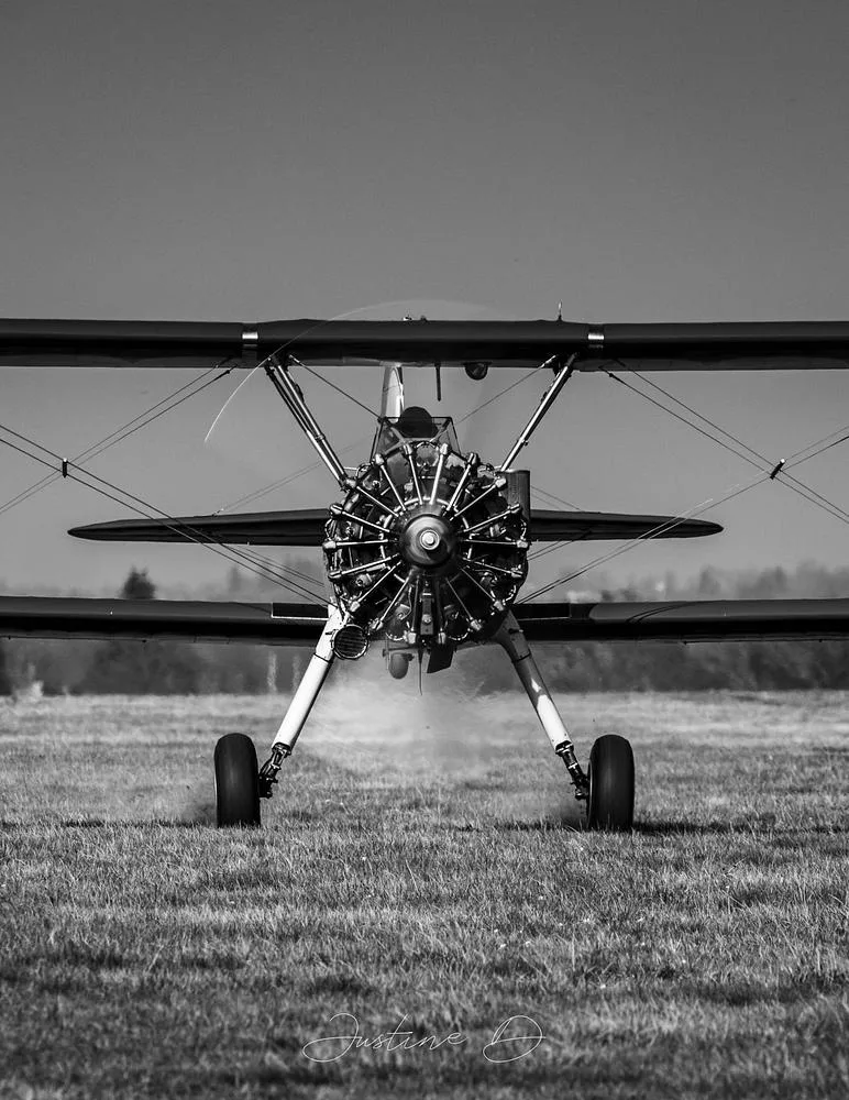 A black and white front view of the biplane, N777CH, on a grassy field with its radial engine running.