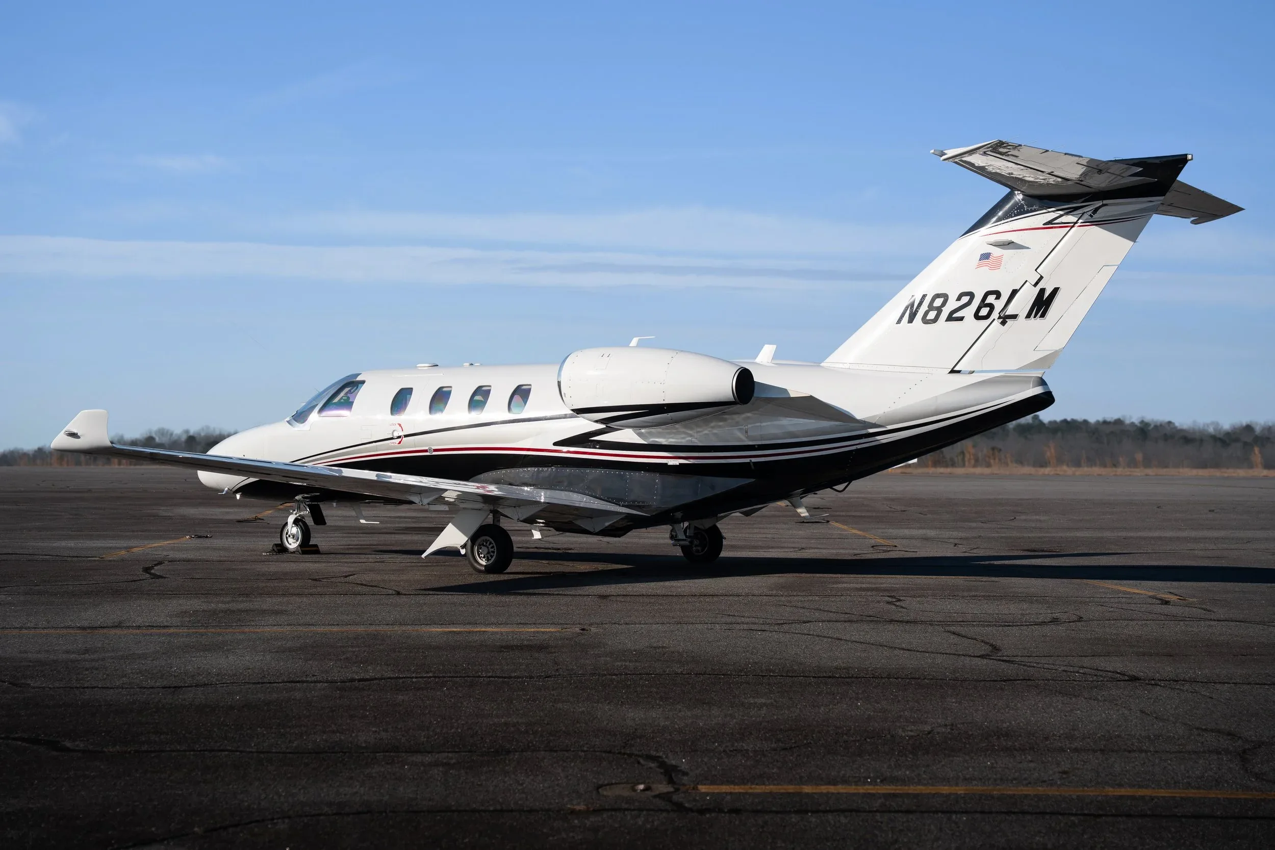A white business jet with black and red stripes, registration N826LM, parked on an asphalt runway under a clear blue sky.