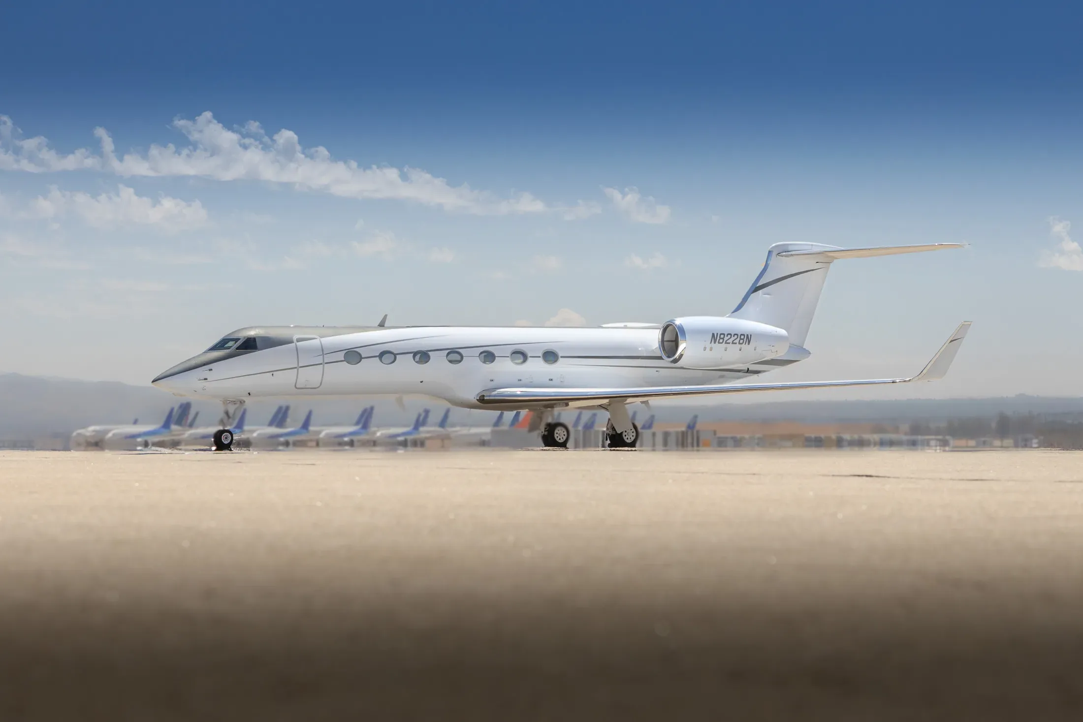 A white Gulfstream business jet with a black nose is parked on an airfield under a blue sky, with other aircraft visible in the background.
