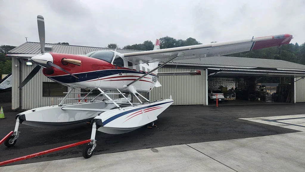A three-quarter front view of a red, white, and blue Quest Kodiak 100 on amphibious floats parked on an asphalt apron.
