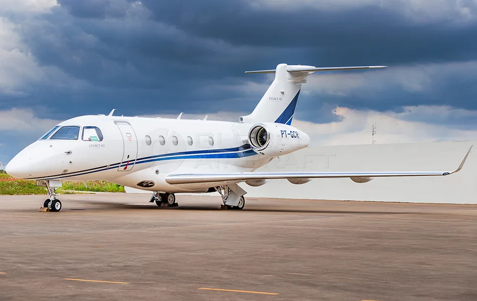 A white Embraer Legacy 450 business jet with blue stripes is parked on an asphalt tarmac under a cloudy sky.
