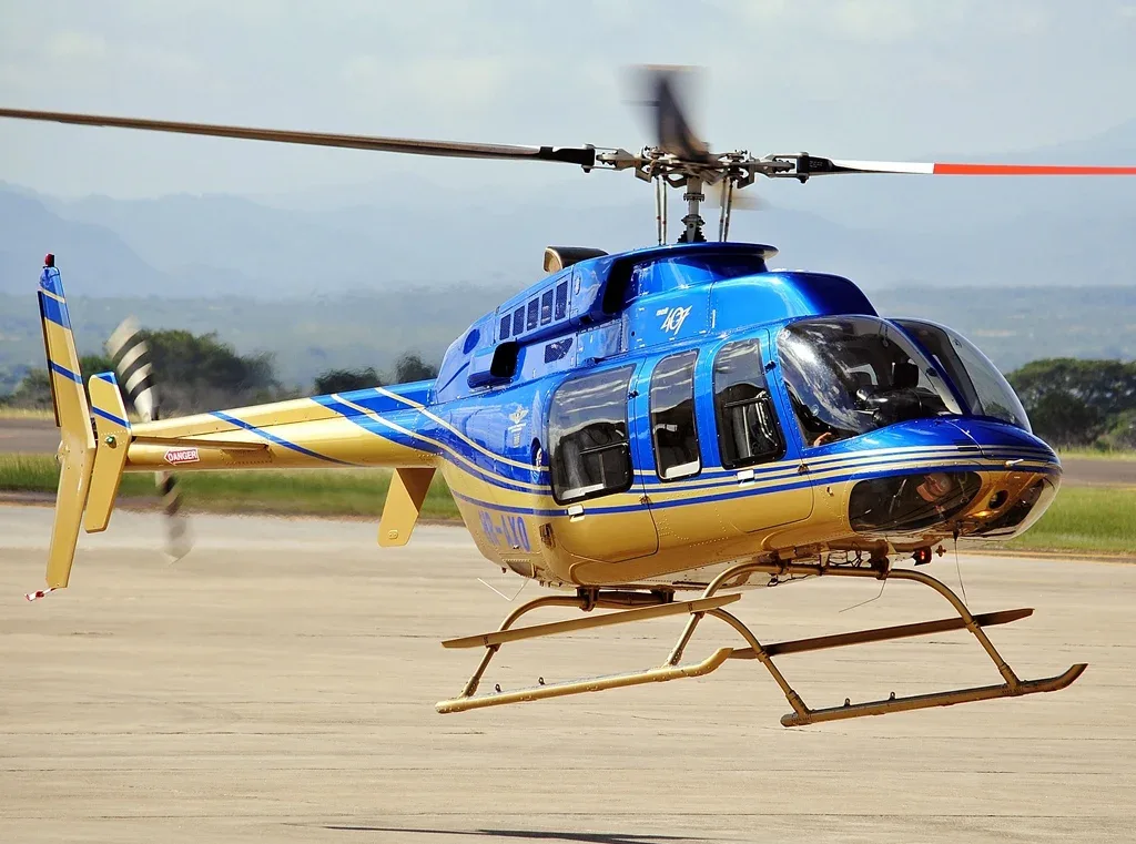 A blue and gold Bell 407 helicopter with registration PR-LXO hovers above the ground on a sunny day.