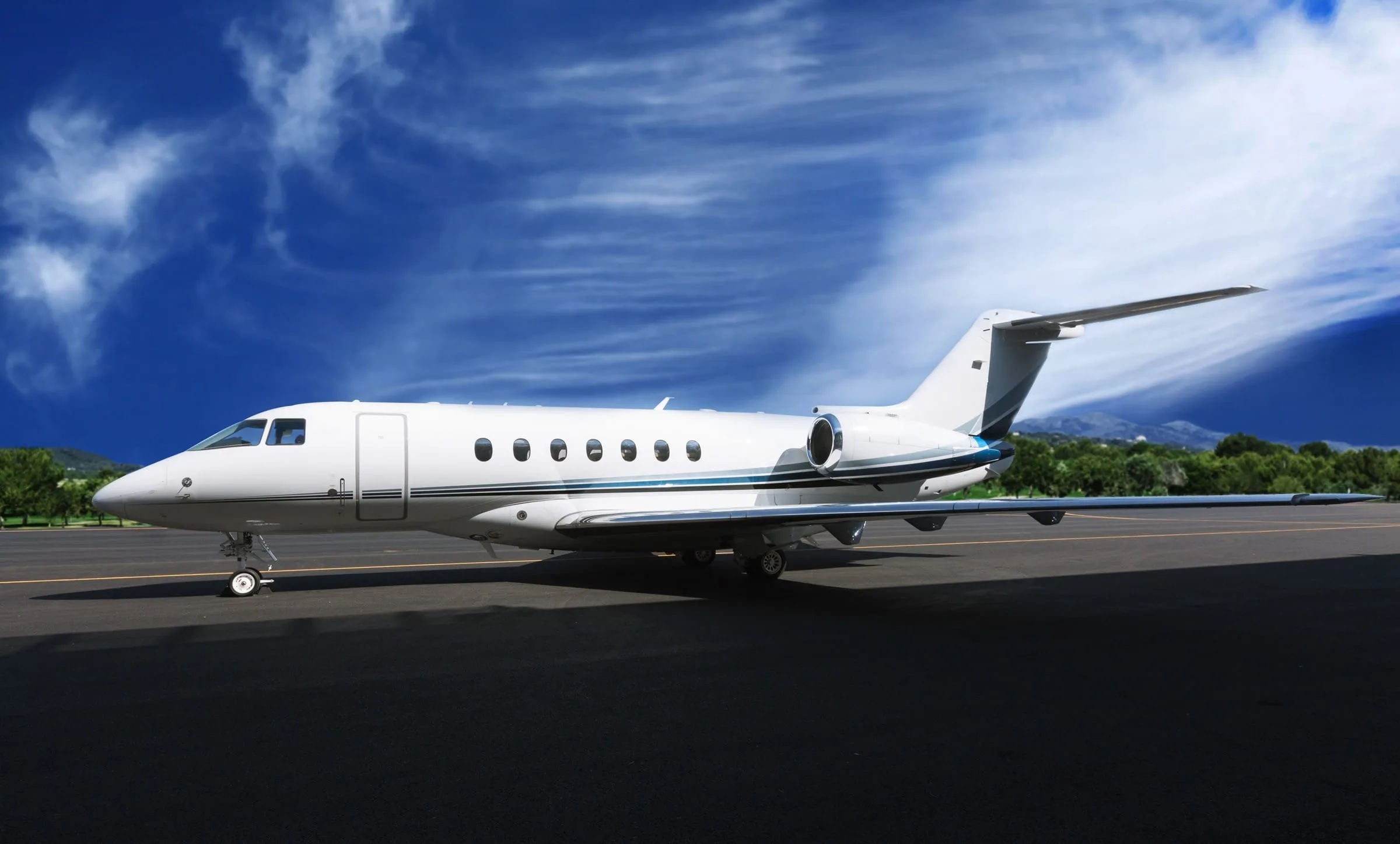 A white private jet with a blue stripe on the tarmac under a blue sky with clouds.