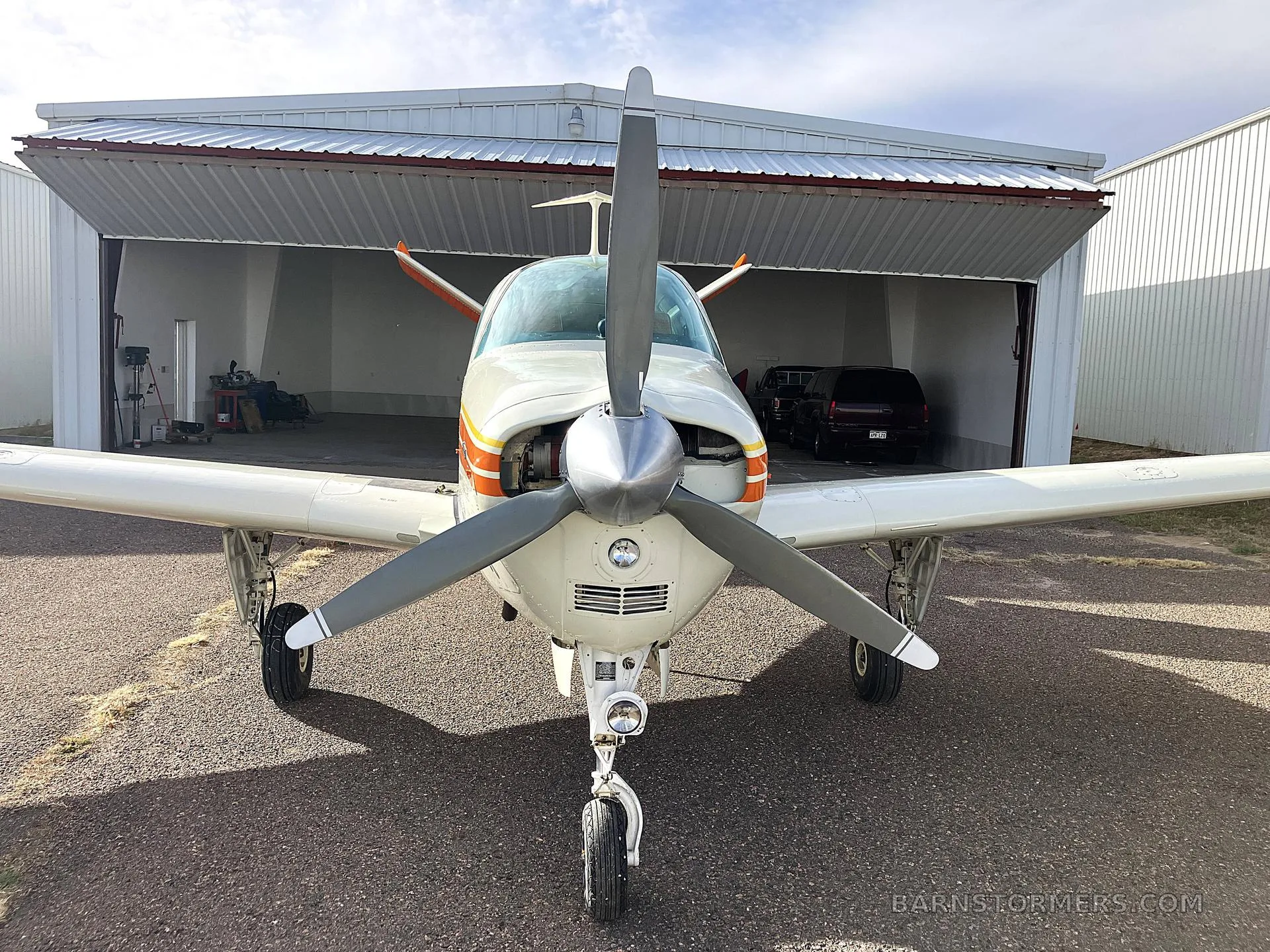 A head-on view of a white, orange, and yellow Beechcraft Bonanza N725AP with its propeller centered, parked in front of an open hangar.