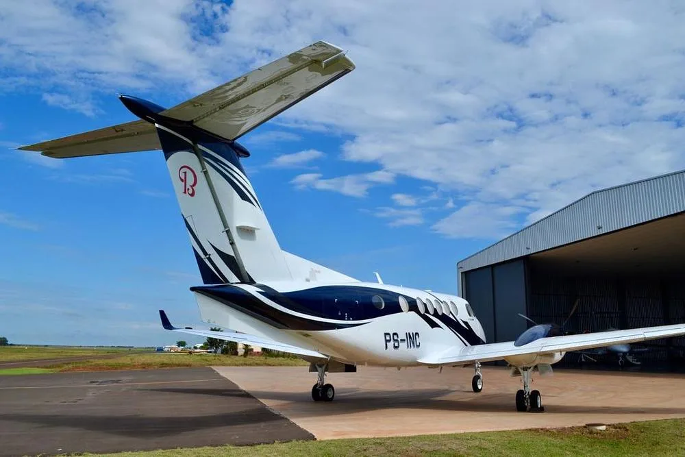 A rear-quarter view of a white and blue Beechcraft King Air twin-turboprop aircraft with registration PS-INC parked on a tarmac next to a hangar.