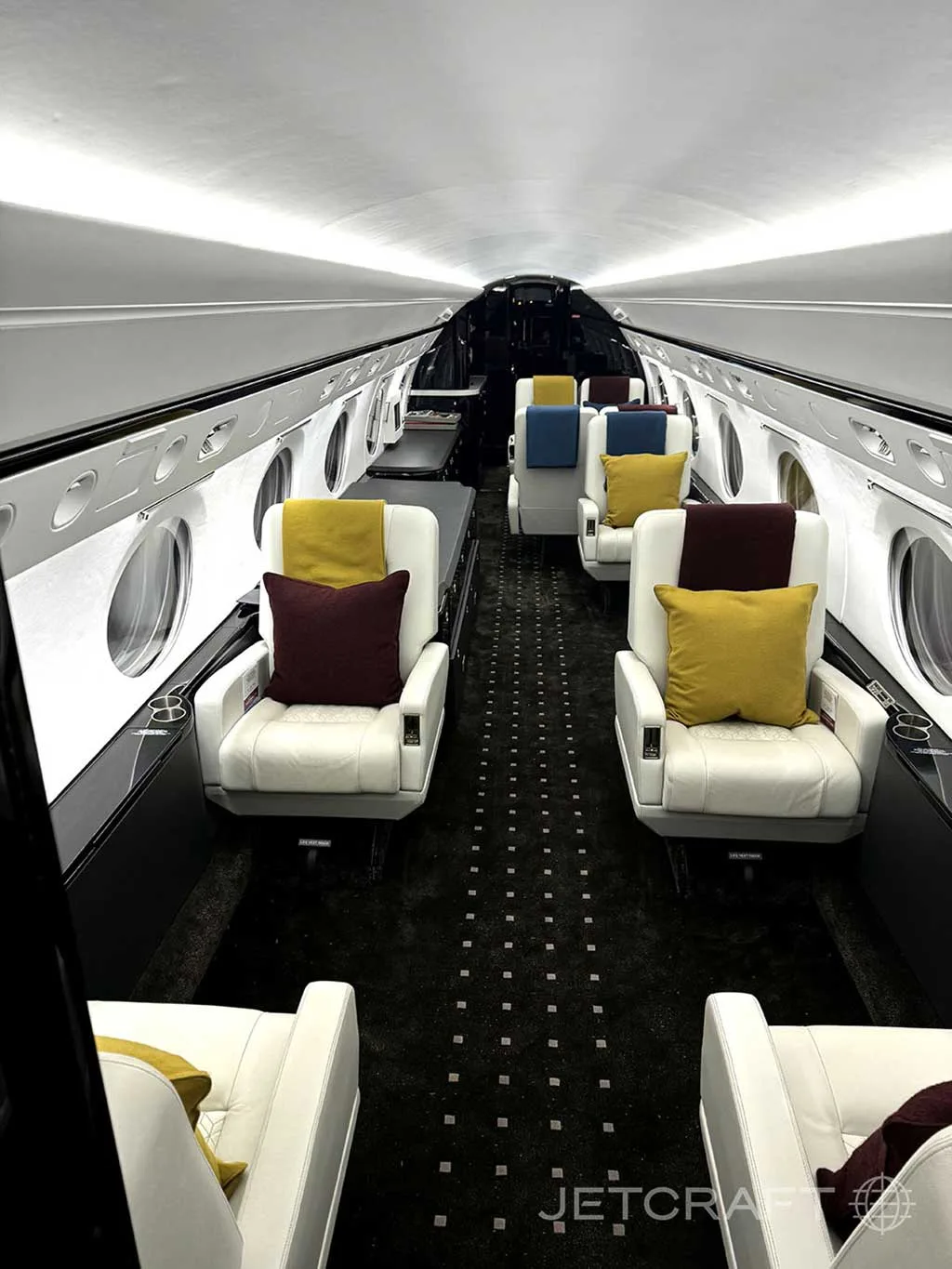 The luxurious interior cabin of a private jet, featuring white leather seats with colorful pillows and a dark patterned carpet.