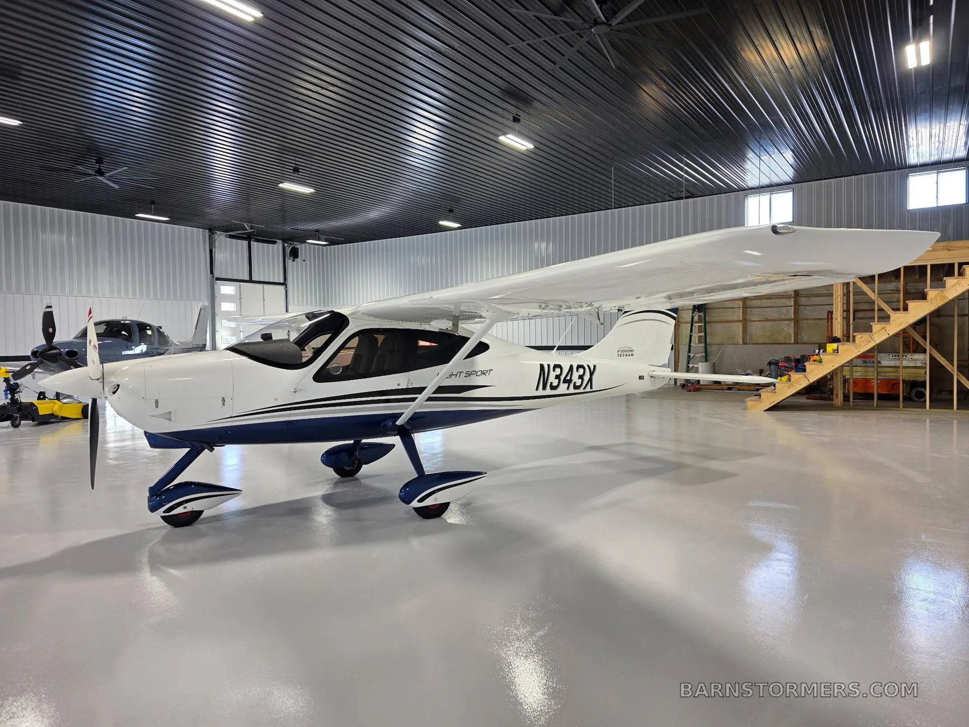 A front-side view of a white Tecnam P2008 Light Sport aircraft with blue accents, registration N343X, parked in a hangar with a wooden staircase in the background.