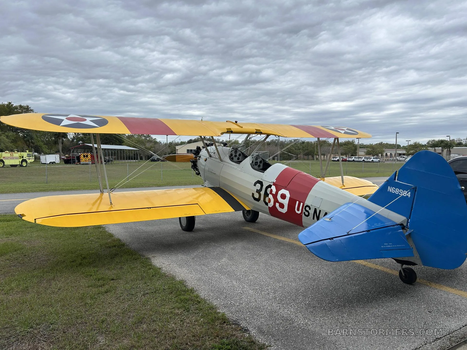 A yellow and white biplane with 'US NAVY 369' markings and a blue tail, seen from the rear-left on an asphalt ramp under a cloudy sky.