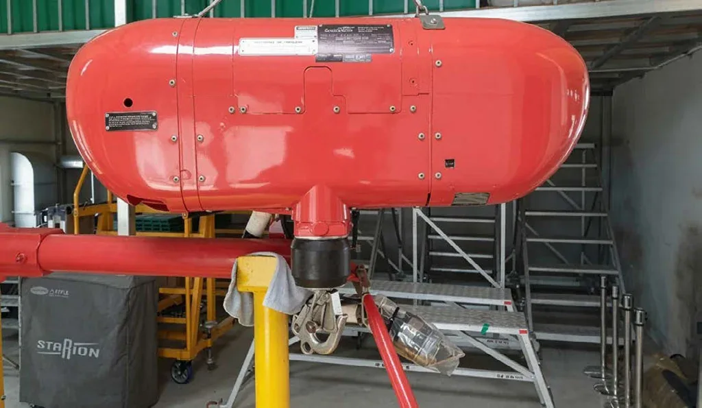 A red external pod or fuel tank for a helicopter is suspended in a workshop setting.