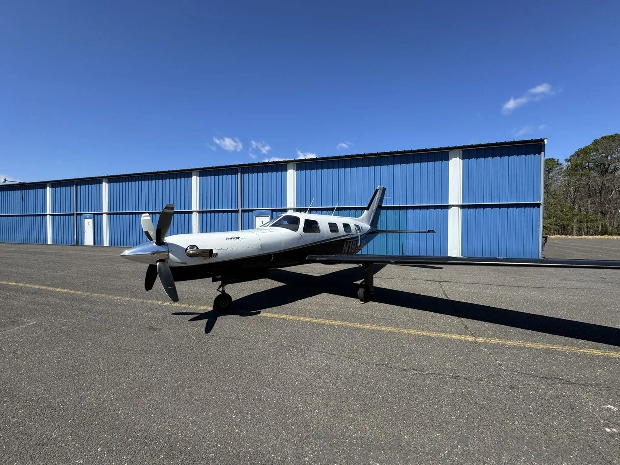 A side view of a white and black Piper M-class aircraft, tail number N195JG, parked on an asphalt ramp next to a blue hangar.