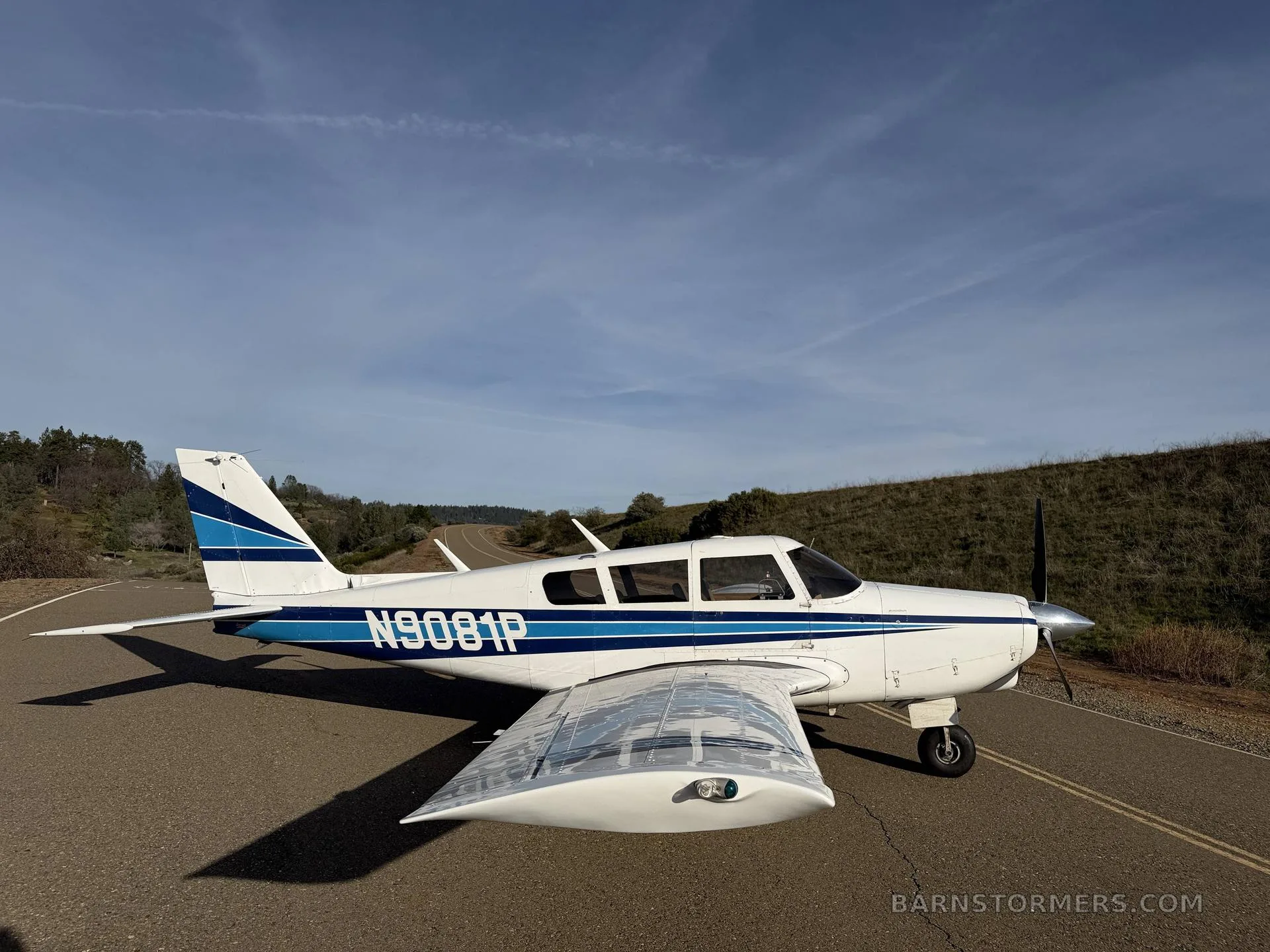 A white single-engine propeller aircraft with blue stripes (N9081P) is parked on an asphalt surface with hills in the background.