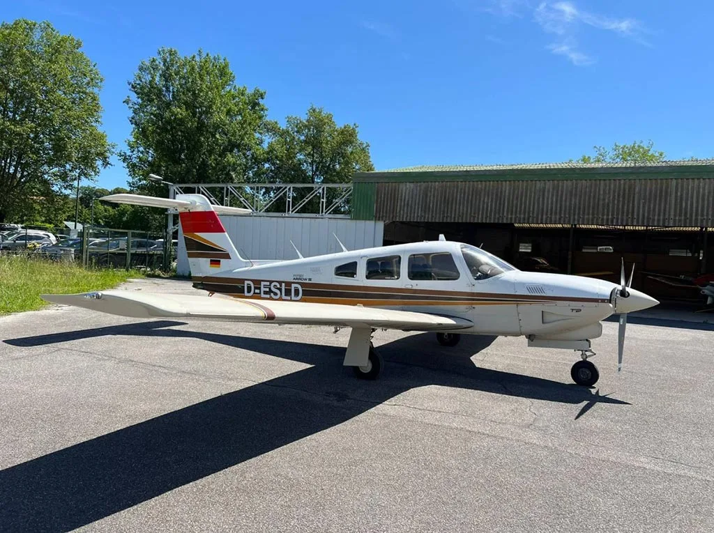A white and brown Piper Arrow III (D-ESLD) parked on an apron next to a green hangar.