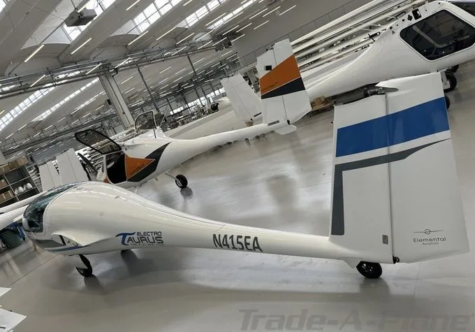 A white Electro Taurus glider with blue and grey tail accents, parked in a well-lit hangar with other aircraft.