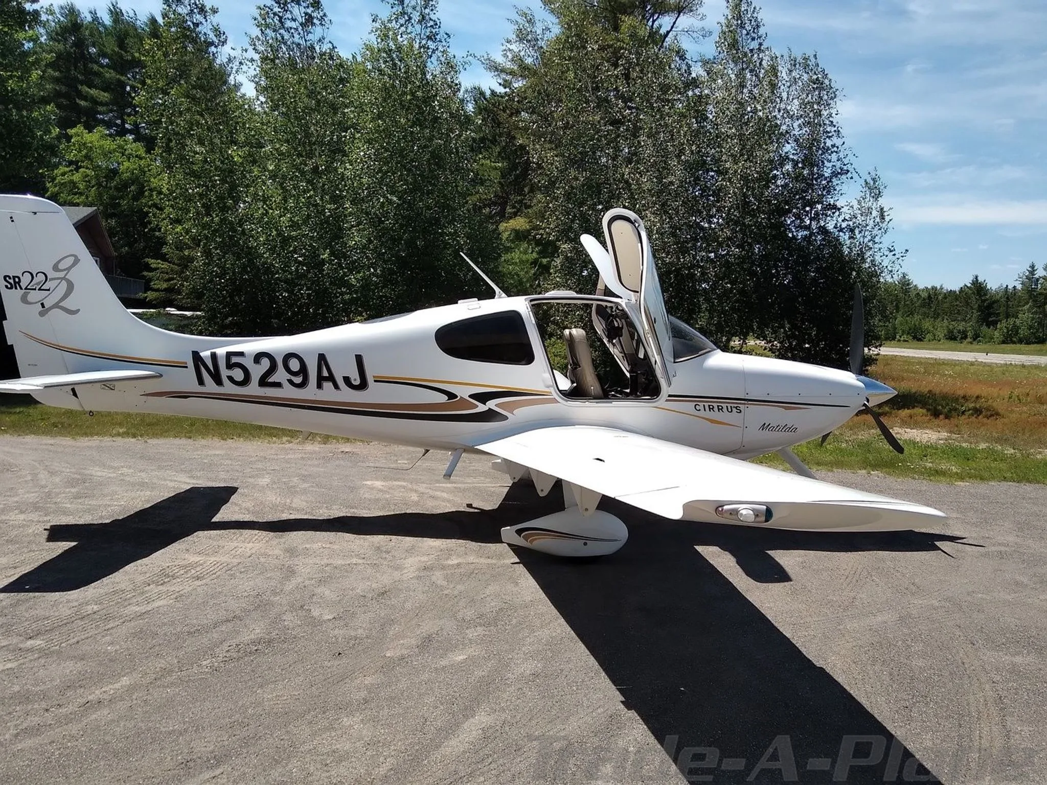 A white Cirrus SR22 aircraft with registration N529AJ and both doors open, parked on a paved surface surrounded by trees.