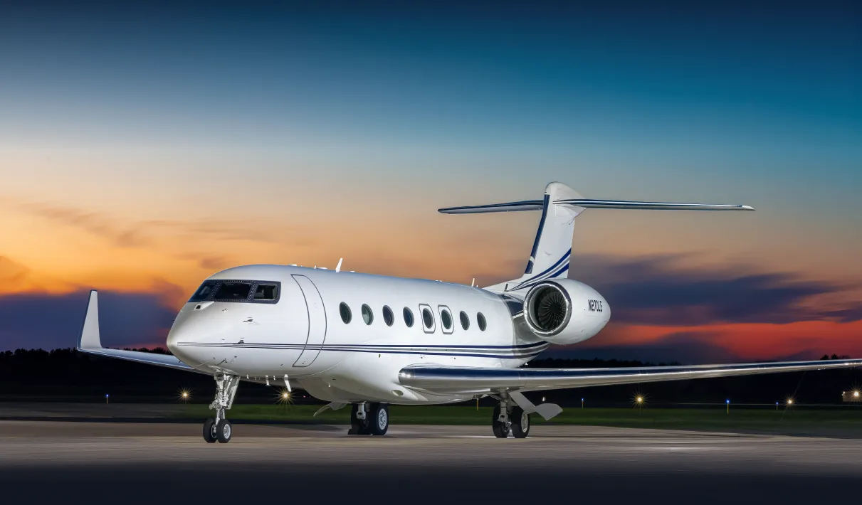 A white Gulfstream G650 business jet with blue stripes is parked on a tarmac at sunset.