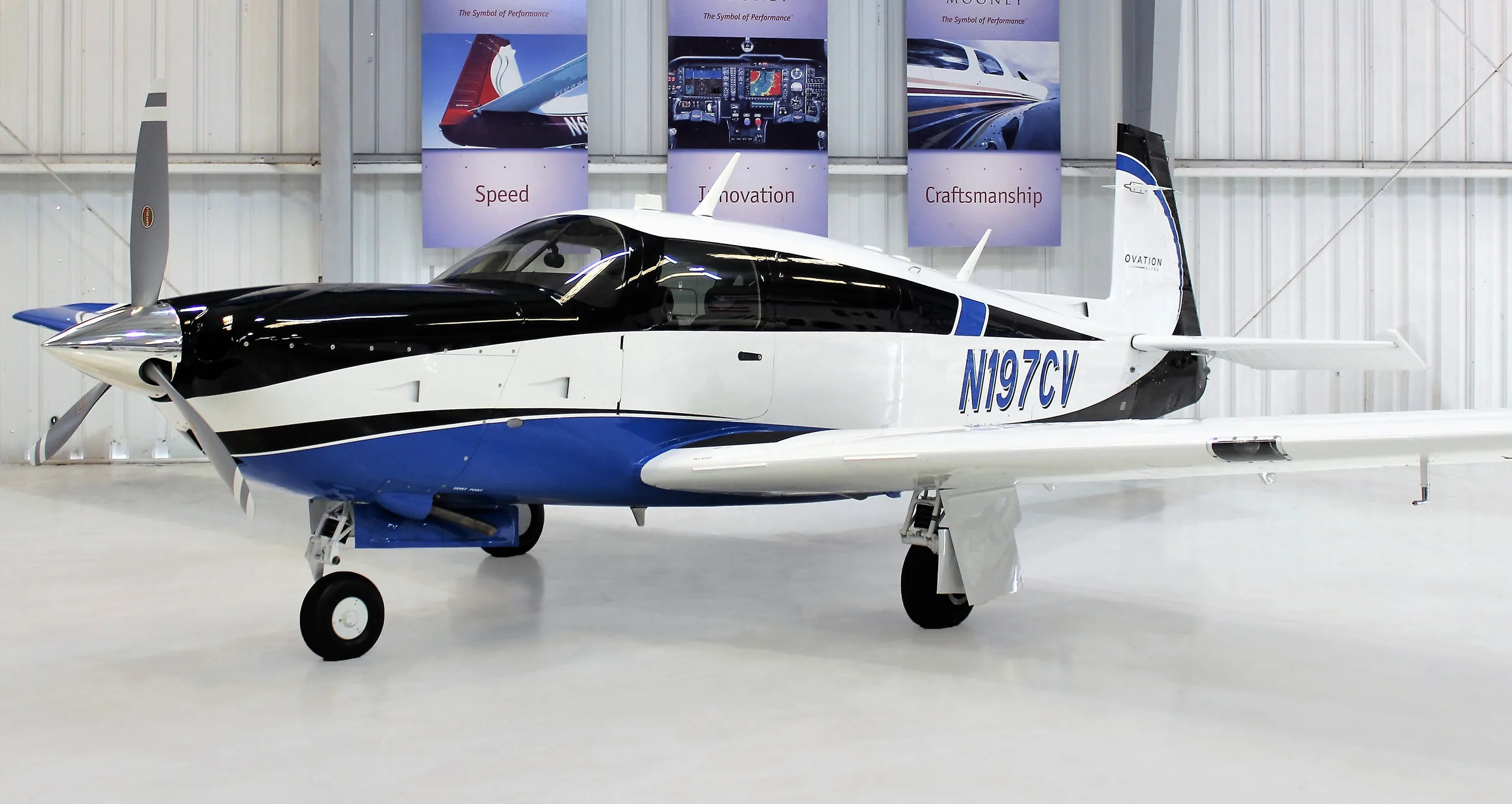 A white, black, and blue Mooney Ovation Ultra aircraft with registration N197CV parked inside a hangar.