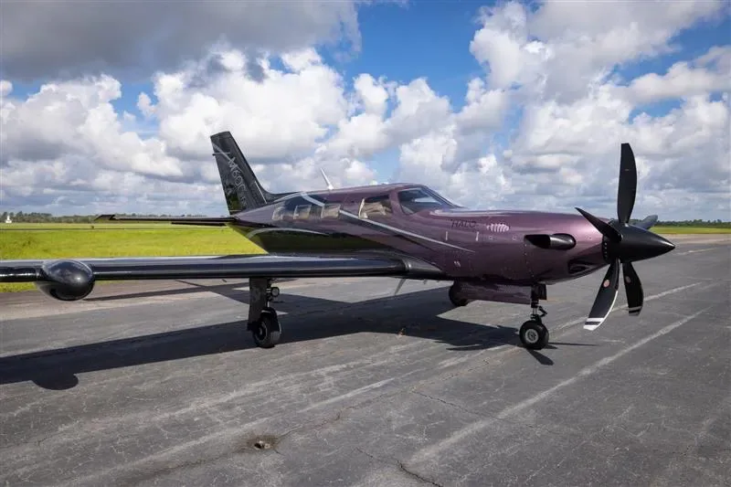 A purple and black Piper M600 turboprop aircraft is parked on a tarmac under a partly cloudy sky.