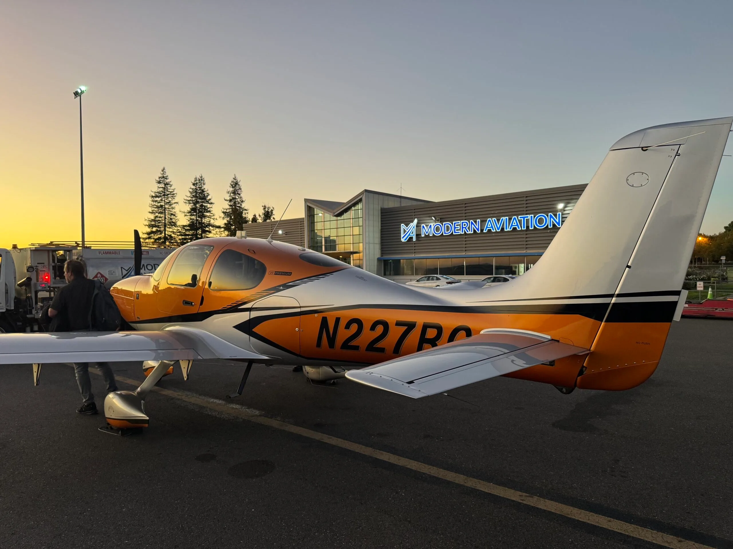 A Cirrus SR22 aircraft with orange and silver paint parked on an tarmac at sunset, with a "Modern Aviation" building in the background.