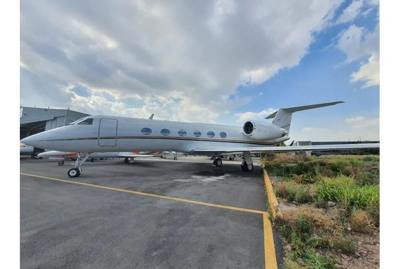A white business jet with a gold stripe parked on an asphalt apron next to a hangar under a partly cloudy sky.