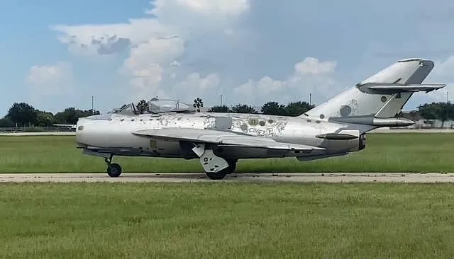 A silver MiG-17 jet aircraft with peeling paint, taxiing on a grassy runway under a cloudy sky.