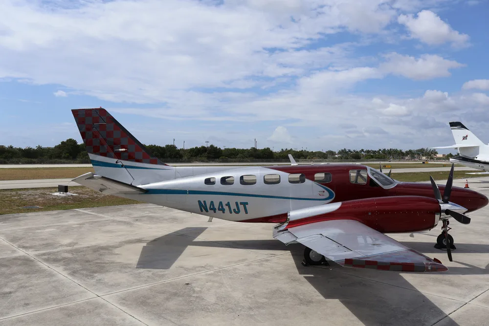 A rear-quarter side view of the red and silver twin-engine aircraft N441JT on an airfield under a partly cloudy sky.