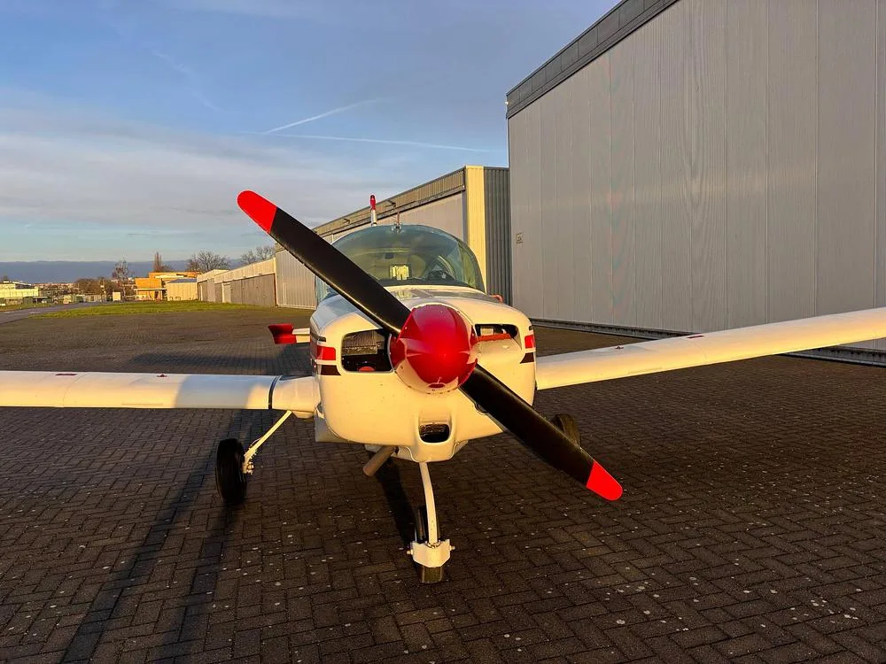 A head-on view of the white Grumman American aircraft, N277MW, showing its propeller with red tips and red spinner.
