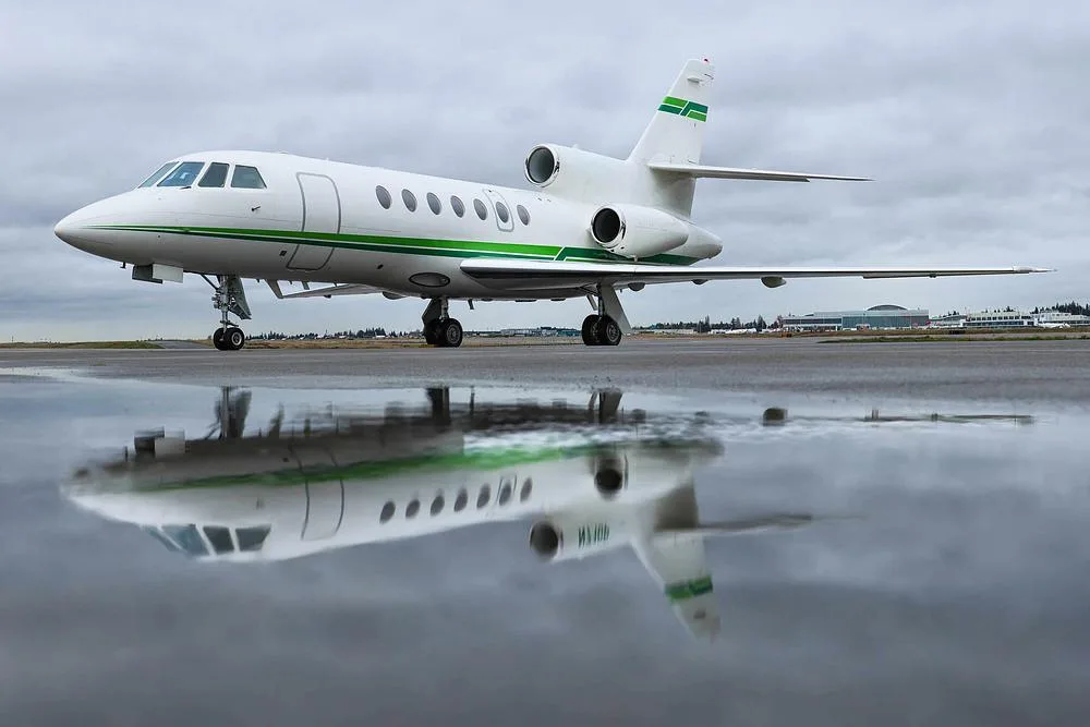 A white Dassault Falcon 900 business jet with green stripes on a wet tarmac, reflecting in puddles under a cloudy sky.
