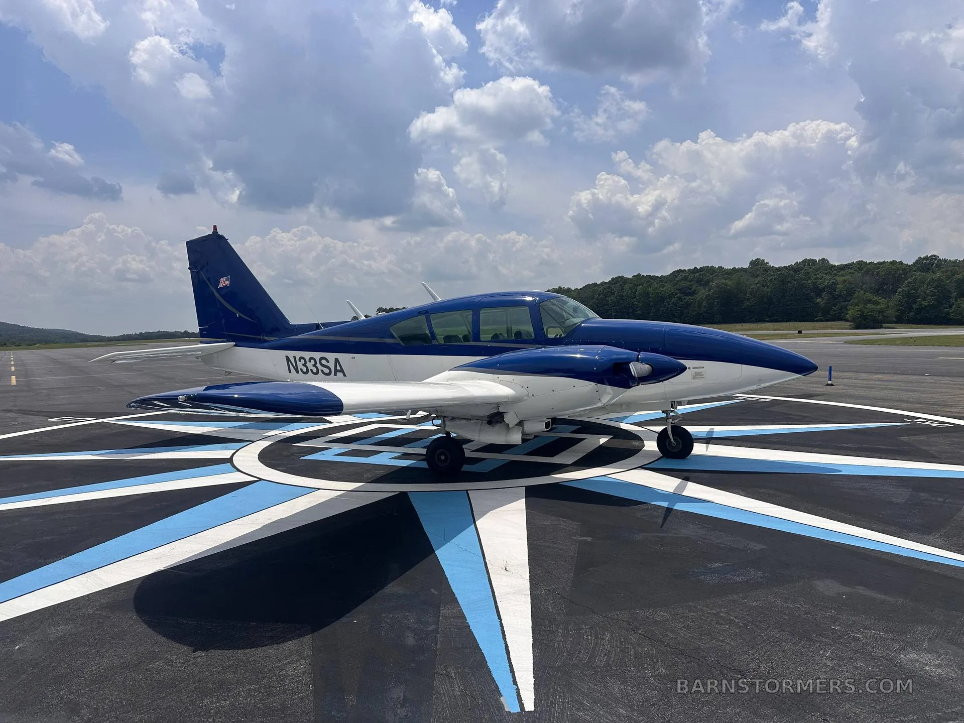 A blue and white single-engine aircraft (N33SA) is parked on a compass rose marking on an airfield under a partly cloudy sky.