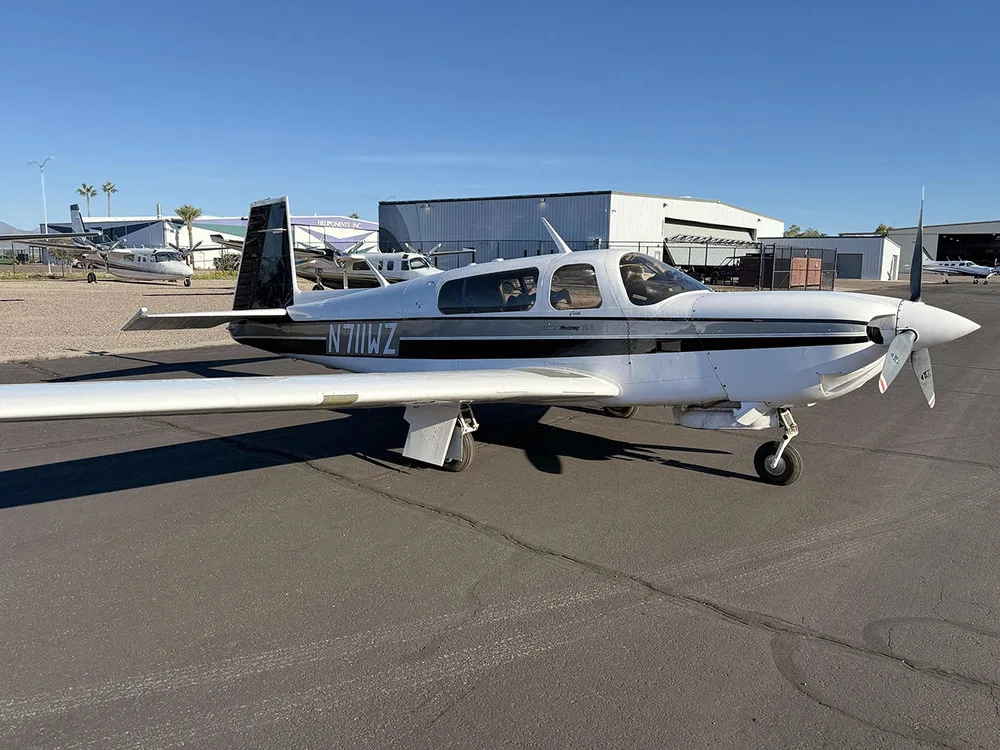 A front-right view of a white Mooney aircraft with black and grey stripes, tail number N711WZ, parked on an airfield with other aircraft in the background.
