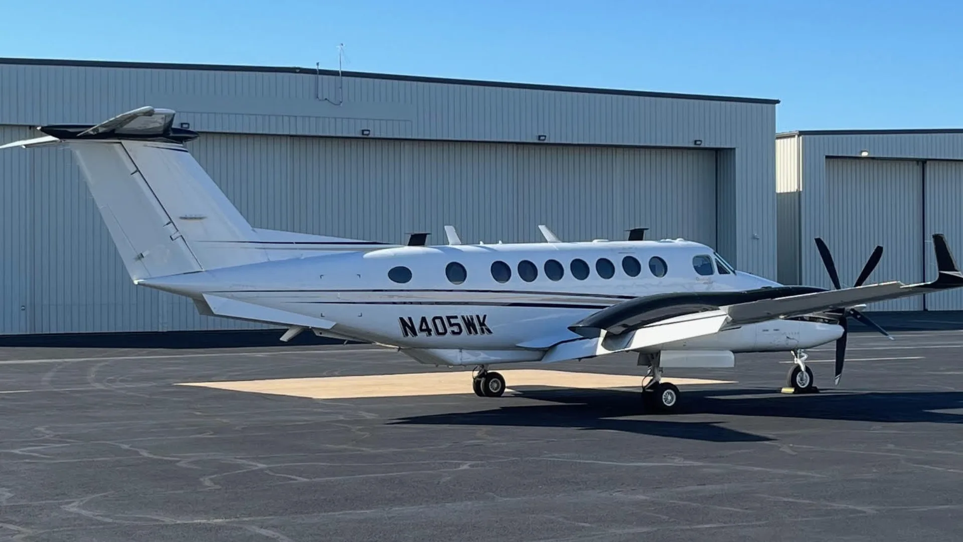 A three-quarter view of a white and black Beechcraft King Air N405WK parked on an asphalt tarmac in front of a white hangar.