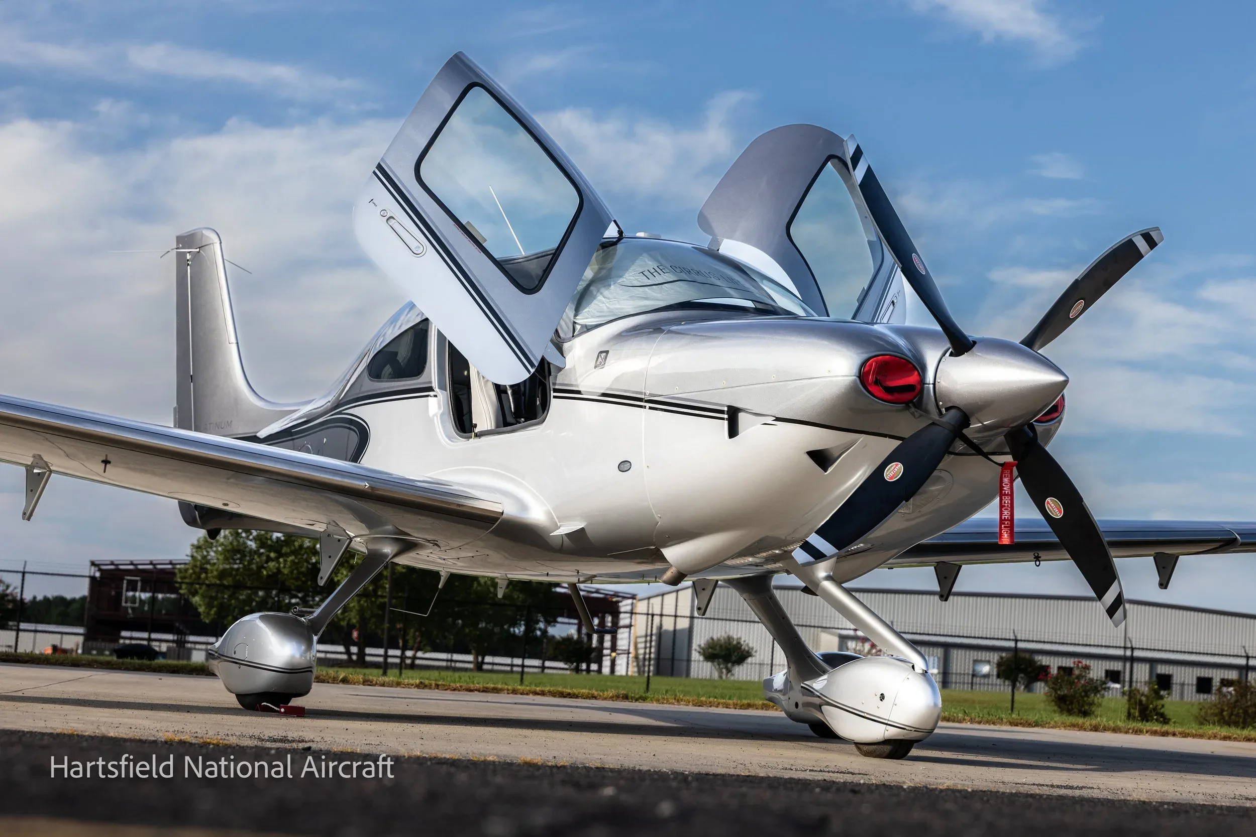 A silver Cirrus SR22 aircraft with its gull-wing doors open, parked on an asphalt surface under a blue sky.