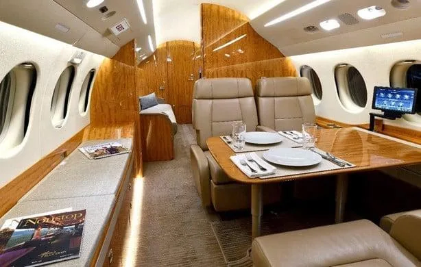 The private jet cabin interior featuring a dining table set for two, a long bench seat, and polished wood paneling.