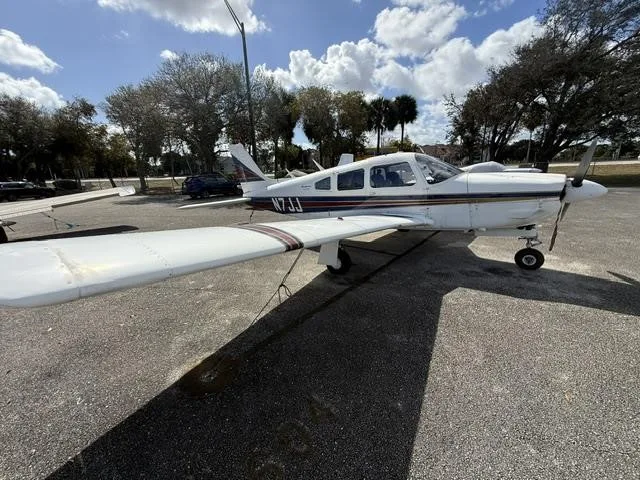 A side view of the white Piper Cherokee aircraft N7JJ with blue and red stripes, parked on an asphalt ramp.