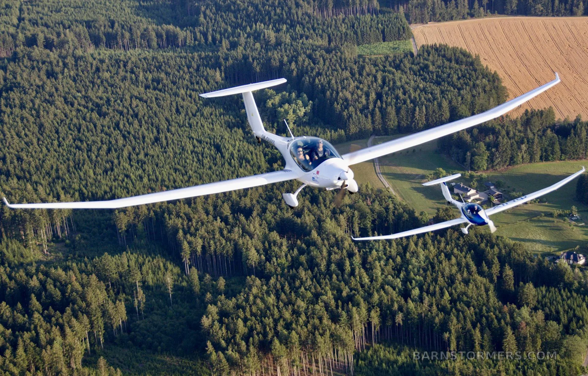 Two white motor gliders fly in formation over a dense forest and agricultural fields.
