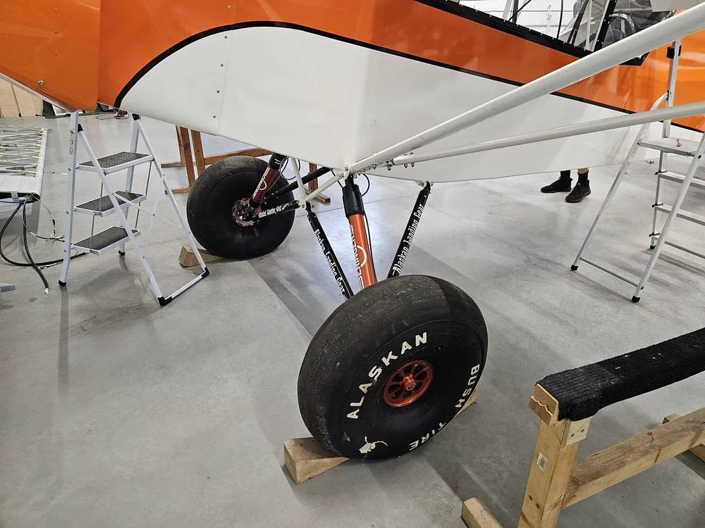 A close-up of the large "Alaskan Bushwheel" tires and landing gear of the orange and white aircraft in a hangar.