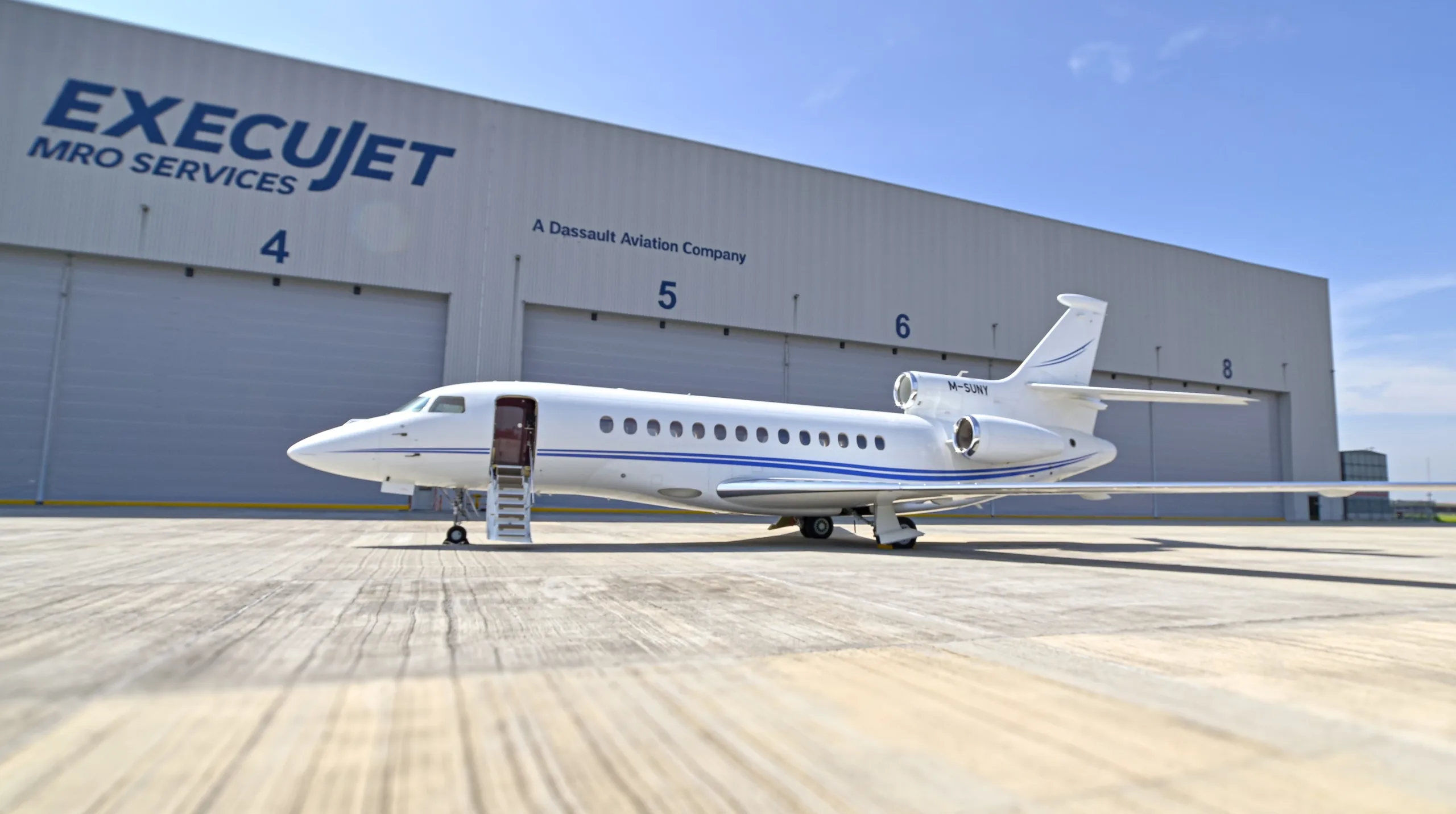 A white Dassault Falcon business jet with blue stripes and registration M-SUNY is parked on an apron in front of an ExecuJet hangar with its main door open.