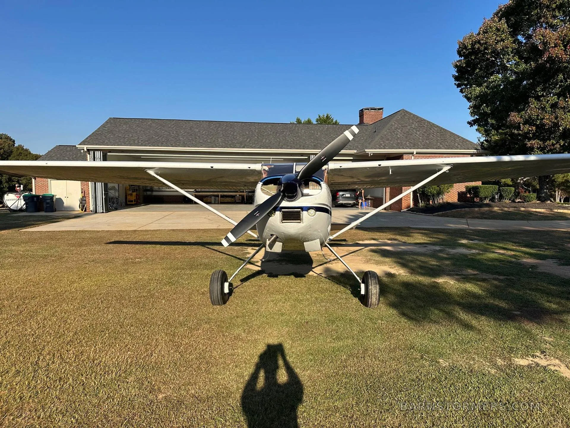 A white and blue Cessna 180 aircraft viewed head-on, parked on a sunny grassy field in front of a hangar.
