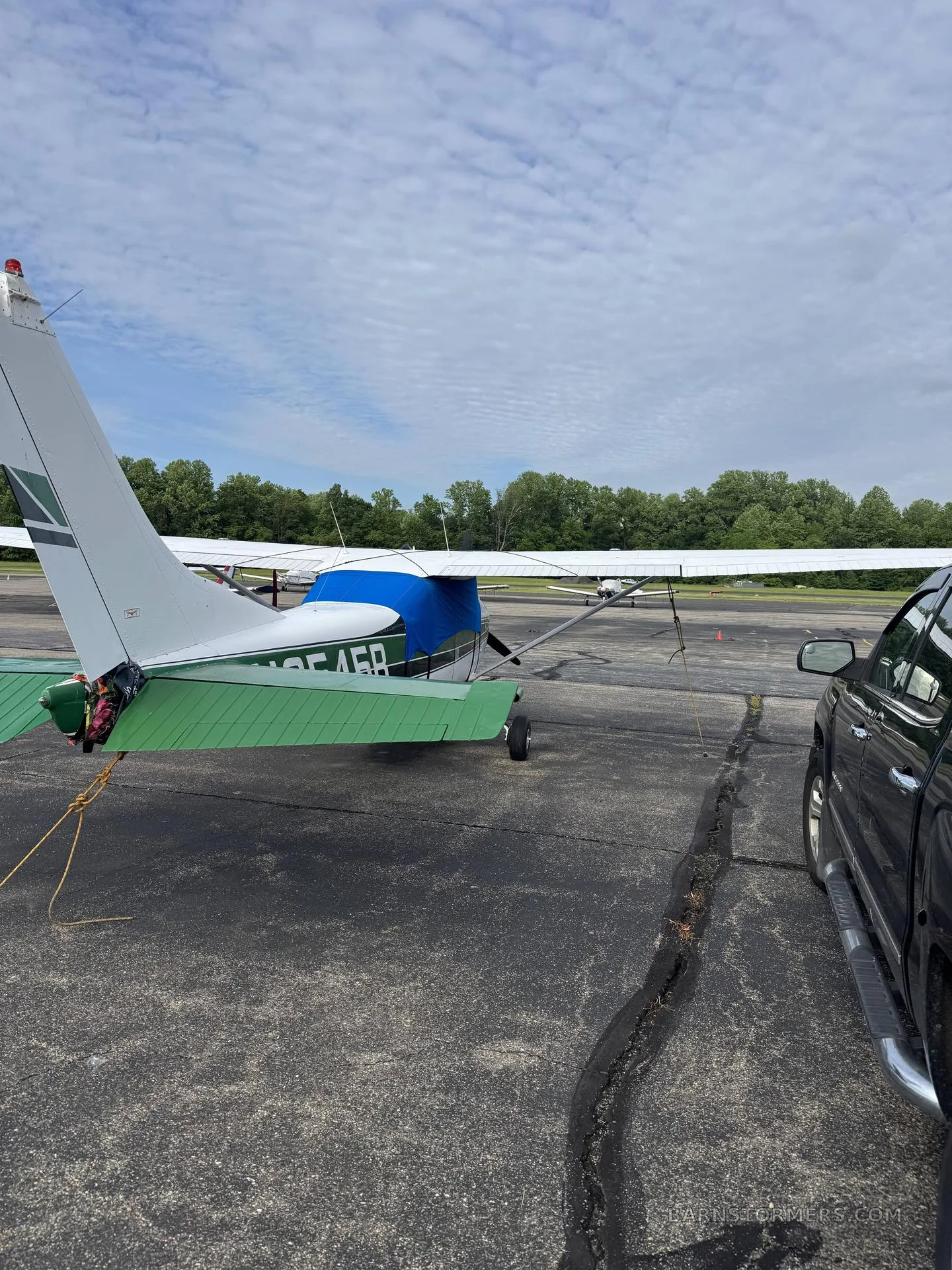 The rear-right view of a white and green single-engine propeller aircraft with a blue engine cover, parked on an asphalt ramp next to a black truck.